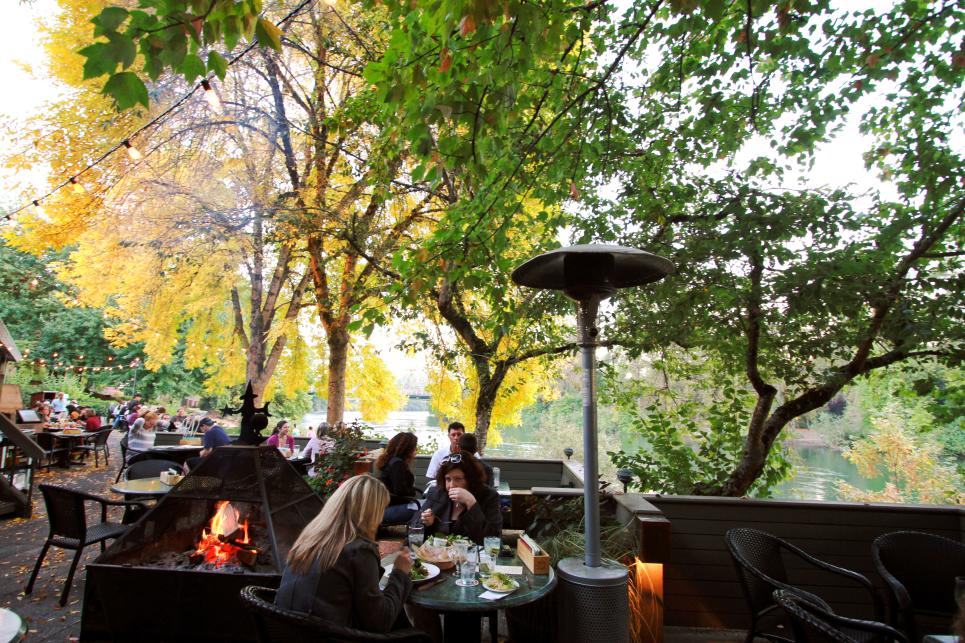 Diners sit at tables by fire pits and under heat lamps on the outside patio. Colorful fall trees and the river are in the background.