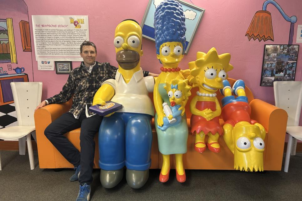 The author's husband sits on the orange couch next to Homer. The whole Simpsons family is crowded onto the couch.