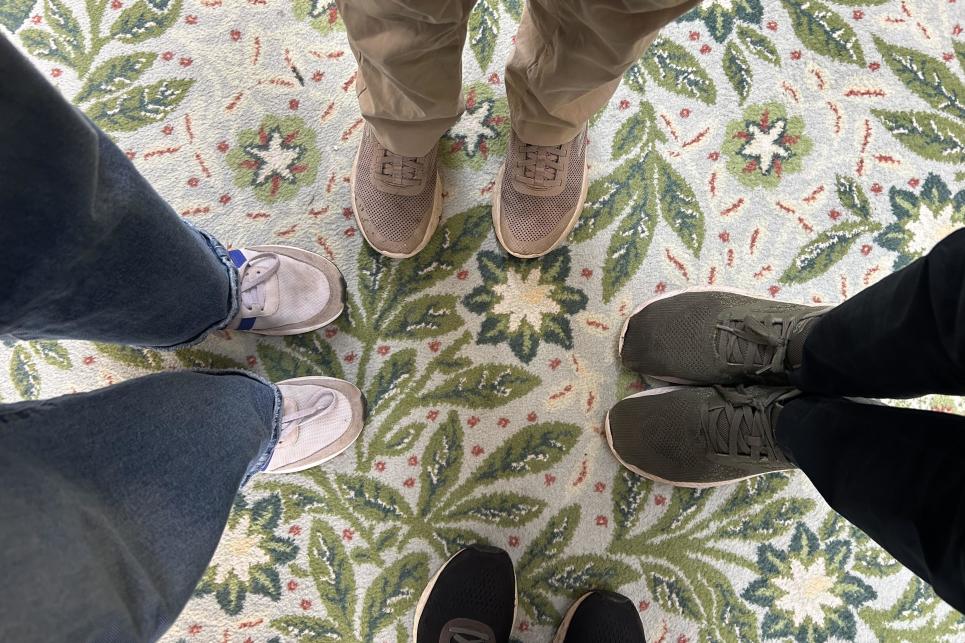 Four pairs of feet standing on the floral leafy green and blue with white flowers and orange speckled patterned carpet.