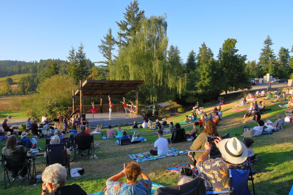 People picnic on the grassy slope around the small wood stage with performers.