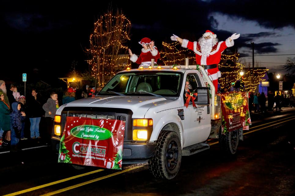 A white truck decorated in twinkle lights rolls down the street with Mr. & Mrs. Claus in the back waving at the crowd.