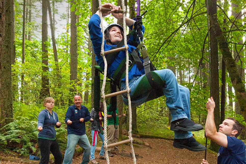 A woman seated in a rope sling uses her arms to climb a rope ladder while a team hoists and stabilizes her assent in a team building exercise on the rope challenge course. The ground is chip bark and big green leafy trees are in the background.