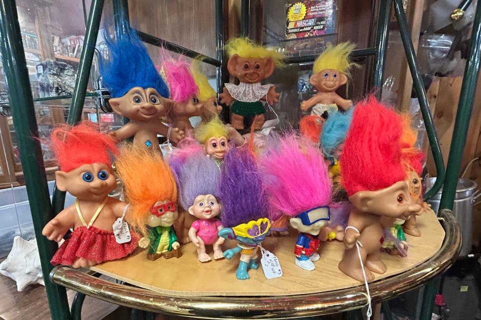 A collection of large and small troll dolls with bright tufts of hair crowd a small shelf.