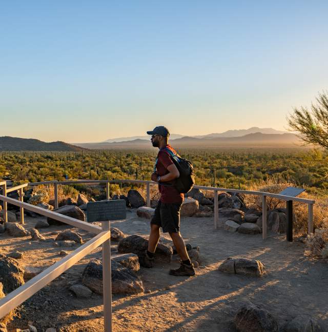Man dressed in boots and hat at a lookout point that outlooks to a desert landscape of brush, cacti, and mountains while hiking in Saguaro National Park