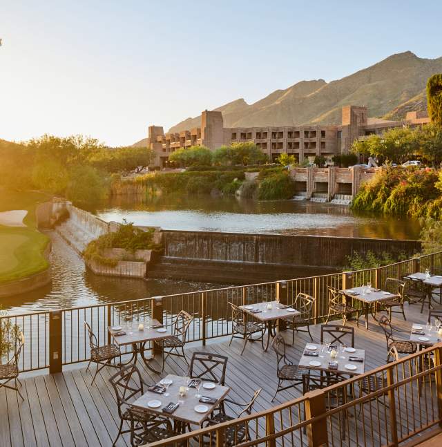 Outdoor dining terrace overlooking a serene water feature and golf course at sunset, with the Loews Ventana Canyon Resort and Santa Catalina Mountains in the background.