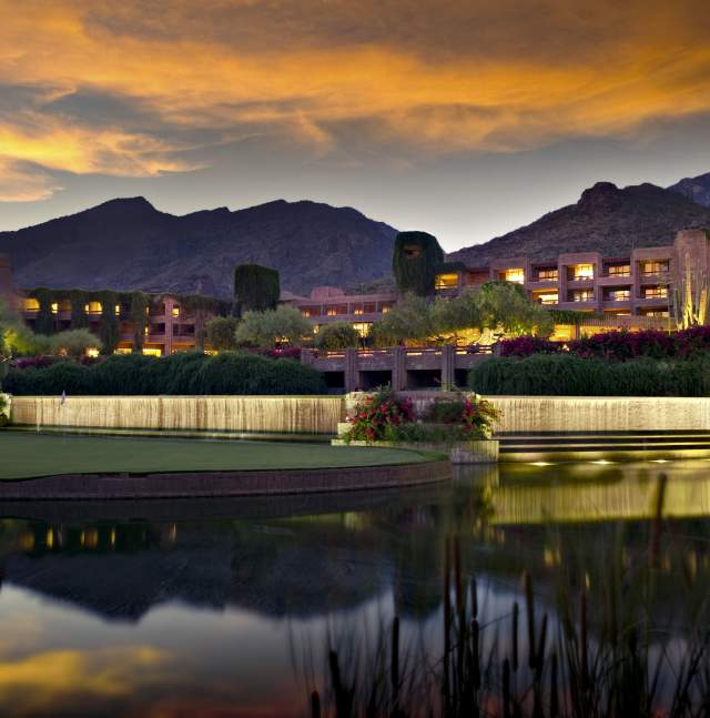 Evening view of Loews Ventana Canyon Resort in Tucson, Arizona, with warm lights glowing from the building nestled against the Santa Catalina Mountains. A tranquil waterfall flows into a reflecting pond in the foreground, surrounded by lush greenery, desert flora, and a softly lit golf course under a dramatic sunset sky.