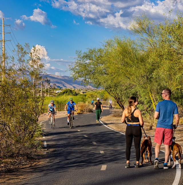 People and dogs walking on paved trail. People riding bikes beside people. All surrounded by greenery