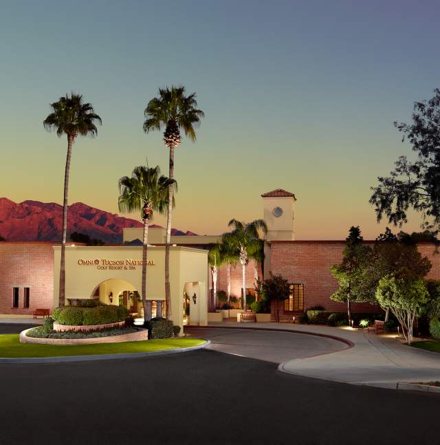 A building with a sign that reads 'Omni Tucson National Golf Resort & Spa' is shown at dusk. The building has a clock tower and is surrounded by palm trees and well-manicured landscaping. In the background, there are mountains under a clear sky transitioning from sunset to twilight