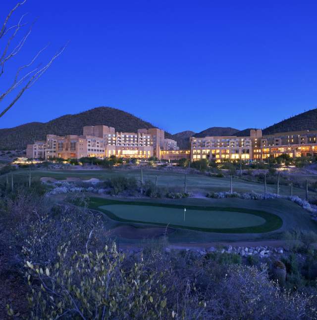 Warm-lit, adobe-style resort nestled in saguaro-studded mountains at dusk, with a manicured golf green and footbridge in the foreground.