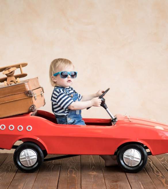 Toddler in child-sized red car, wearing sunglasses with suitcases
