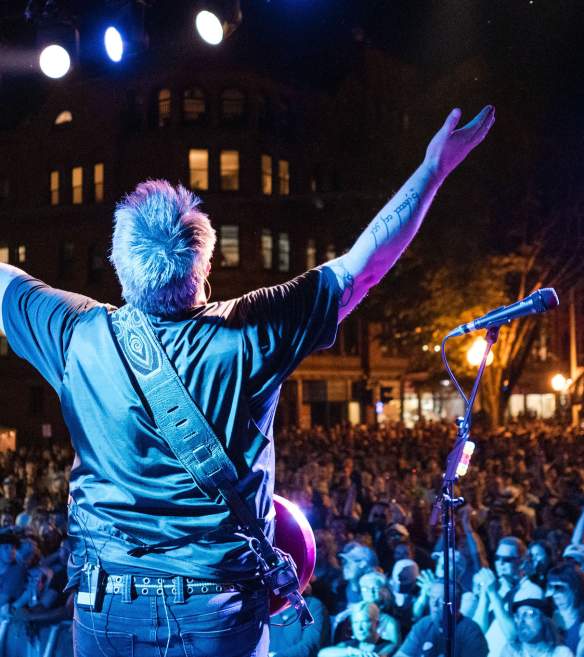 guitar player on stage with hands up in front of huge crowd at night