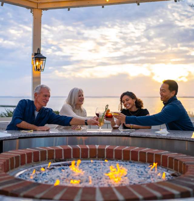 Four people gather around a circular fire pit, toasting drinks as the sun sets over a serene waterfront. Lanterns illuminate the scene.