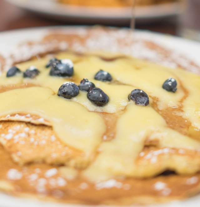 A close-up of a plate of pancakes topped with blueberries and syrup, with a blurred figure in the background pouring syrup. Glasses of orange juice are visible.
