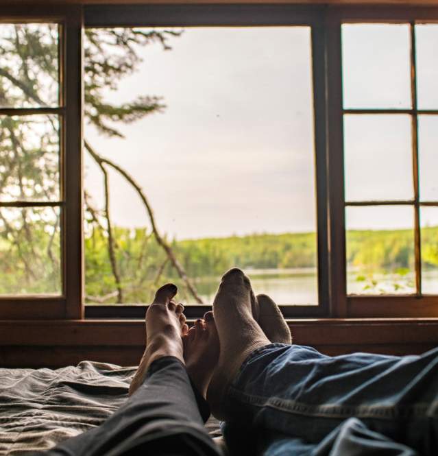 Two people's feet with the window showing view on the lake/wilderness from their cabin
