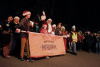 Santa Claus leads the Candlelight Walk 2024 along Washington Avenue, walking in front of a group of participants carrying a festive banner.