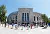 Daytime straight-on view of a Yankees Stadium entrance with a plaza full of fans