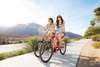Two women biking down sidewalk with mountains in the background.