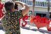 Man in patterned shirt taking a photograph of a woman in an orange dress posing among large red heart sculptures outdoors.