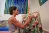 A woman in a tank top and apron paints flowers on a canvas. Other paintings hang on a white wall behind her.