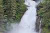 frothy white waterfall cascades through evergreen covered mountains onto the rocky seawead covered beach at low tide and into the bright green ocean