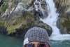 frothy white waterfall cascades down a rocky mountain into the green sea. Woman smile for selfie in front wearing a bright orange Mustang life suit, pink tinted aviator glasses and a black and white fitted beanie over strands of blonde hair