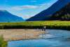 A little girl squats as Dad and son stand fishing on the River Rock embankment stretches into a steady streaming slough of blue waters that flows around and beyond tall green seagrass as dramatic tall jagged mountains "V" into the ocean in the distance under blue skies with white scatter clouds.