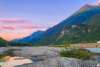 A stone covered riverbed strewn with fallen trees and stumps lies in between two tributaries of aqua colored water; one flowing rapidly and the other, so still that it reflects the blue, purple and pink sunset sky lying overhead. Giant jagged dark green and blue mountains rise beyond the sky scape on the right with one rolling green tree covered hill to the left.