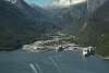 Layers of steep towering mountains from snow capped to evergreen covered mountains dive into a narrow valley with a river on the left and three cruise ships positioned  at fingerlike dock infrastructure