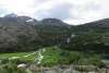 wide angle view of waterfall rolling over a massive mountain of rolling rock hills that give way to a lush green valley floor where the falls fork out into multiple streams and into a lake off to the right under overcast skies