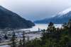 View of the Taiya inlet from Dyea Rd. Overlook shows Skagway valley buildings, docks, trees and an Alaska Marine Transport Lynden barge on the calm water under overcast skies