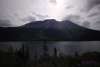 Cloudy skies lit up by the Sun trying to escape from behind a dark blue/black mountain that looms over the lake in a light rain. Evergreen trees at the forefront