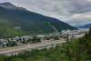 Vantage point of Skagway from the Lookout on Dyea Road show a long narrow airstrip and townsite nestled perfectly in the valley floor with giant evergreen covered mountains steeply jetting up behind it. Three Cruise Ships sit iddle on calm dark seas.