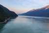 The wooden rail of a cruise ship deck is barely visible in the left corner of the photo that gives way to lightly roughed up dark blue green waters between two steep evergreen covered jagged mountain ranges in North America's deepest fjord under pink and blue sunrise skies