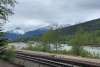 chain link fence in front of railroad tracks with Skagway River flowing in the background and green trees and blue snow capped jagged mountain peaks in the distance under grey cloudy skies