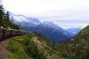 A line of vintage train cars curves along the mountainside, cutting through forested and rocky terrain. Snow-capped peaks rise over the White Pass and Yukon Route in Skagway.