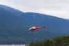 A red helicopter flies over Skagway’s rocky shoreline and forest, with mountains rising in the background under a gray sky. The light is soft, bringing out the textures in the trees and the turquoise water.