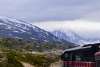A train on the White Pass and Yukon Route curves along the track through rugged terrain in Skagway, Alaska. Snow-covered mountains rise in the background under cloudy skies.