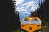 A yellow and green locomotive from the White Pass and Yukon Route drives between pines with snow-covered mountains behind. The train lights shine under an overcast sky, contrasting with the Skagway landscape.