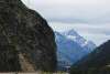 A narrow railway track hugs a rocky cliff edge near Skagway, Alaska, with a sign marking "American Shed." Snow-capped mountains rise in the distance under overcast skies.