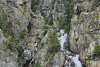 Rocky jagged mountains with scattered green trees on the green water's edge with white frothy waterfall cascading down into the ice cold green sea. Blue sky overhead