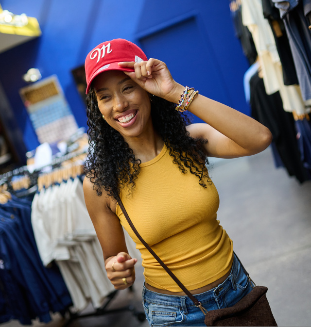 Smiling shopper trying on a red Miller-branded baseball cap inside the Miller Brewery gift shop, surrounded by racks of apparel and branded merchandise.