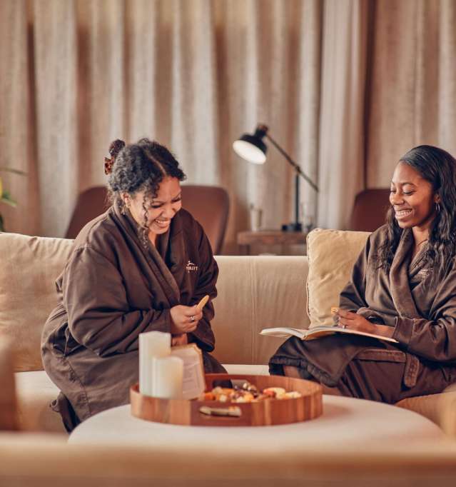 Two people sitting in robes in a spa, laughing and holding beverages.