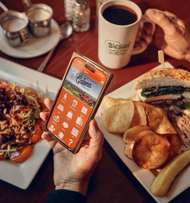 Two people sitting at a table with two plates of food and cups of coffee. One person is holding a phone with the Galena Country app showing.