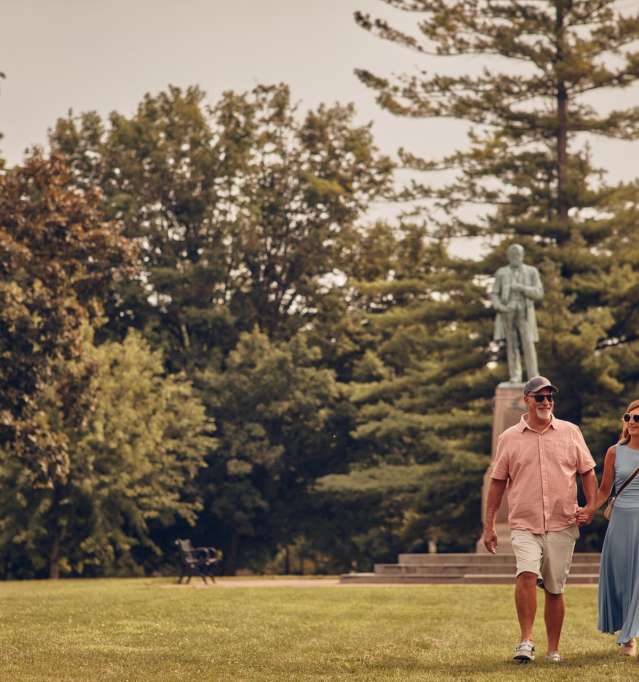 Couple walking in a park with a statue and trees behind them.