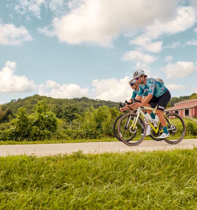Two people riding bicycles on a road with a red barn behind them.