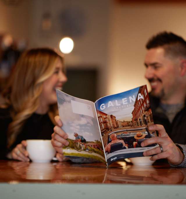 Two people sitting in a coffee shop, drinking out of mugs and holding a Galena Country Visitor's Guide.