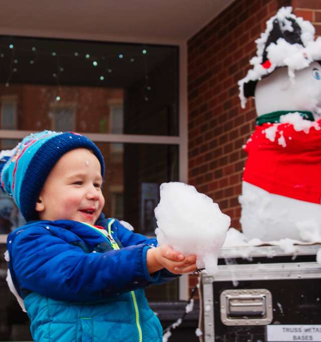 Child holding snow, standing outside a brick building.