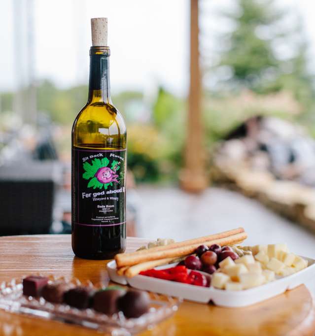 On a table sits a bottle of wine, a plate of chocolates, and a fruit and cheese plate.