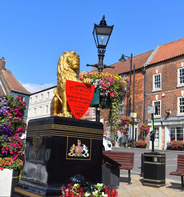 Golden lion water pump on a black plinth with a red commemorative shield, surrounded by colourful flower displays, set in a Caistor Market Place, with red-brick buildings and a lamppost under a clear blue sky.
