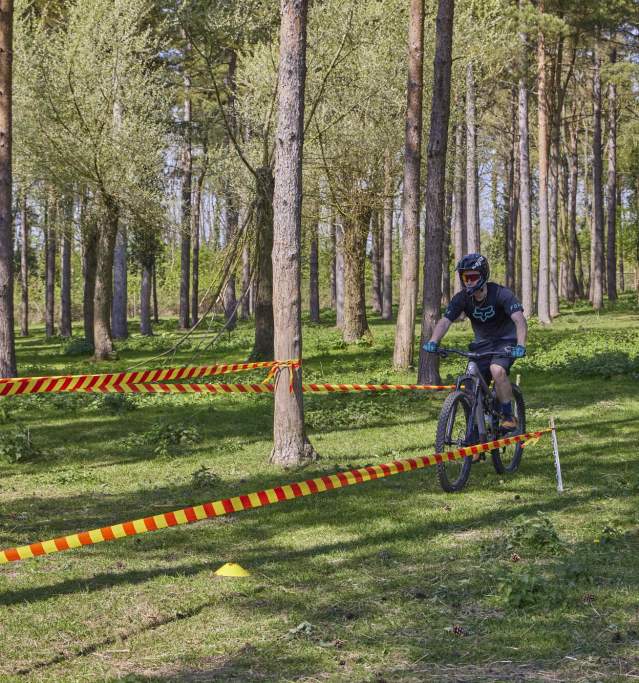 A young man wearing a helmet and goggles rides a mountain bike along a forest trail in the Lincolnshire Wolds, following a marked course lined with red and yellow tape between the trees.